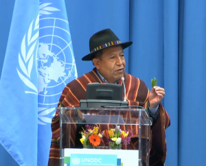 David Choquehuanca Céspedes, Vice President of the Plurinational State of Bolivia. He is standing behind a lectern and addressing the 68th Session of the Commission on Narcotic Drugs in Vienna, Austria. In one hand, he is holding up a few coca leaves.