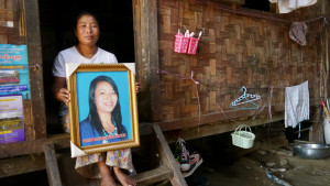 Woman in doorway of woven wood house, holding framed picture of her murdered daughter