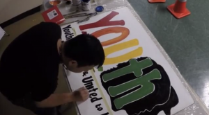 YOUTH Program Grows with Community Center young man painting a sign that says YOUTH in rainbow letters